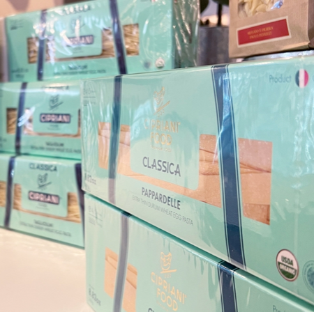 Boxes of Cipriani Food Classica Pappardelle pasta are stacked on display. The packaging is light blue with clear windows showing the pasta inside and features labels indicating it is organic.