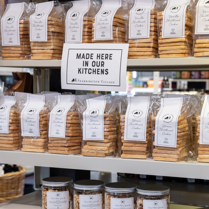 Packages of shortbread cookies are displayed on shelves beside a sign that reads “Made Here In Our Kitchens” at Fearrington Village. Glass jars of food items are visible on a lower shelf.