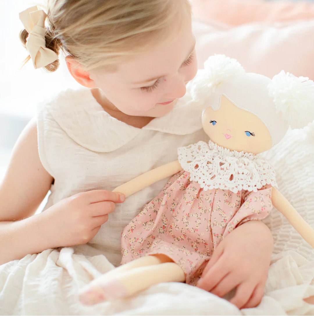 A young girl in a white dress with a bow in her hair smiles while holding a handmade cloth doll dressed in a pink floral dress and a white crocheted collar, sitting on a soft, light-colored blanket.
