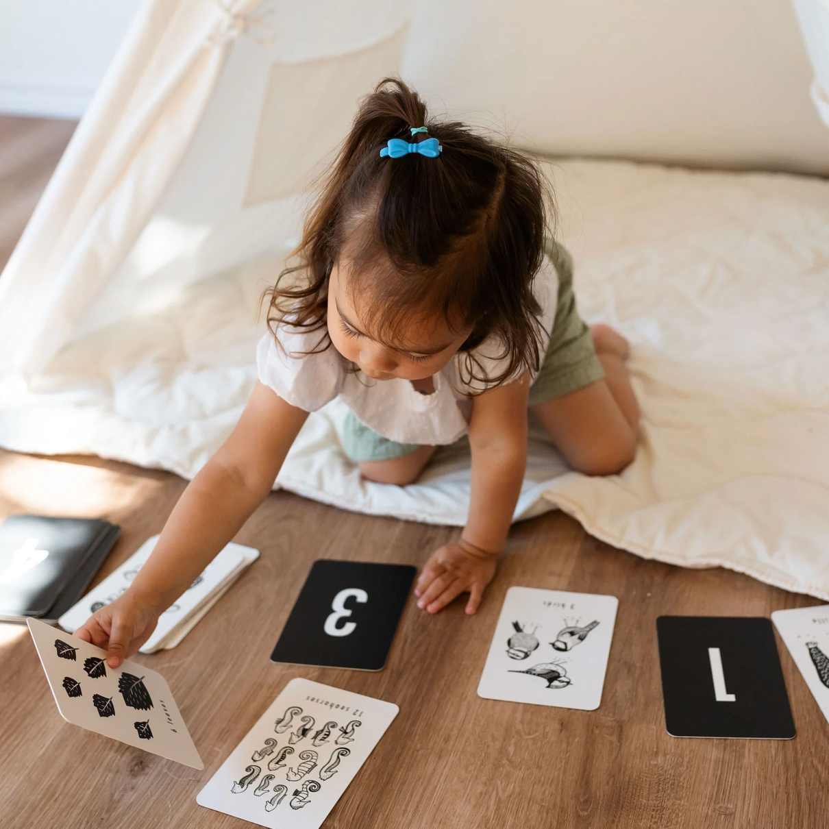 A toddler with a blue bow in her hair kneels on a blanket inside a tent, reaching for a black-and-white flashcard among several cards with numbers and illustrations spread out on the floor.