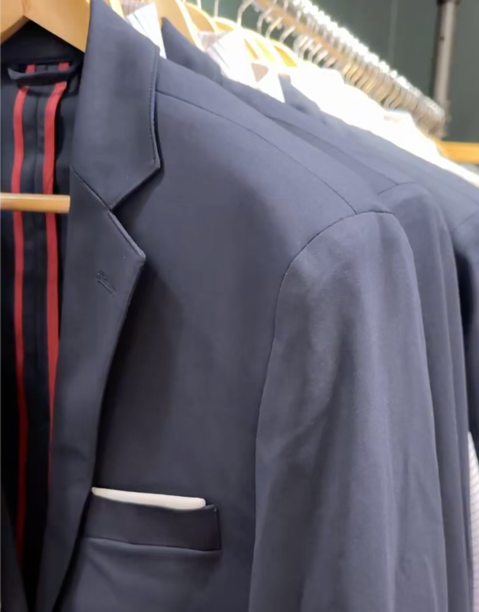 A close-up of navy blue suit jackets hanging on wooden hangers, with a visible white pocket square in the front jacket and red-striped lining inside.