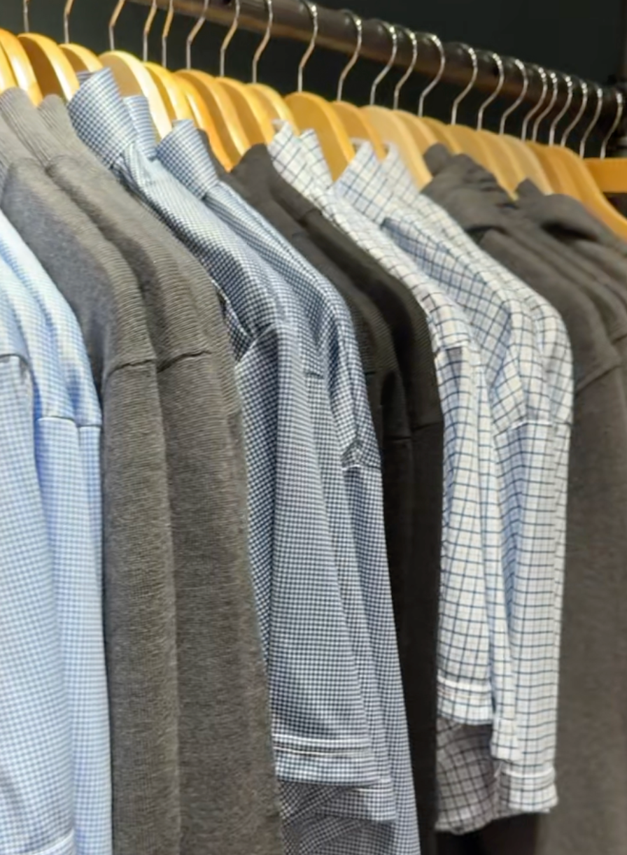 A row of shirts on wooden hangers, featuring a mix of light blue checkered dress shirts and dark gray T-shirts, all hanging on a metal clothing rack.