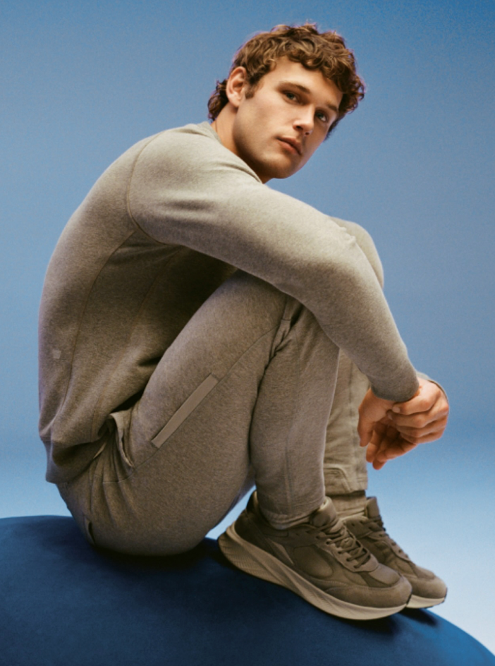 A person in a fitted gray tracksuit and gray sneakers sits on a blue surface against a blue background, looking slightly upward with a relaxed expression.