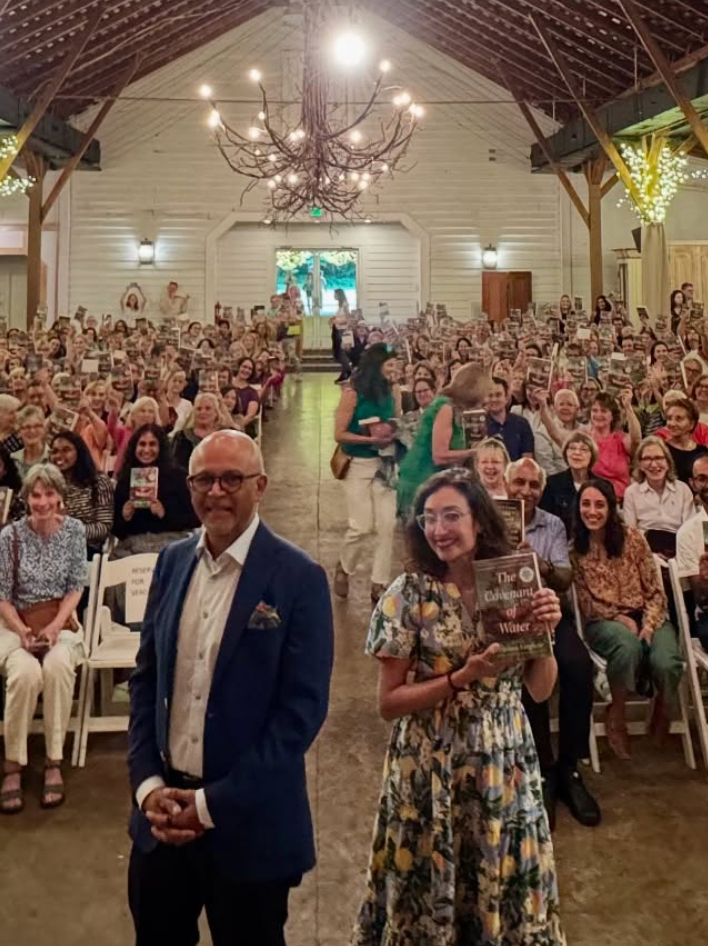 A smiling man in a suit and a woman holding a book stand at the front of a large, brightly lit hall filled with seated people. The atmosphere is cheerful and festive, with string lights and chandeliers overhead.