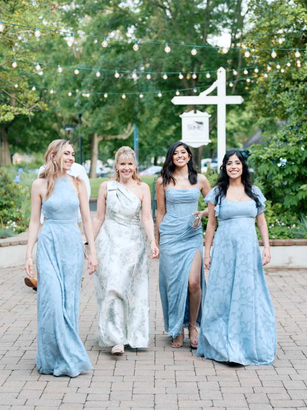 Four women in light blue and white dresses walk together outdoors on a paved path, smiling and talking. Trees and string lights create a festive, garden-like atmosphere in the background.