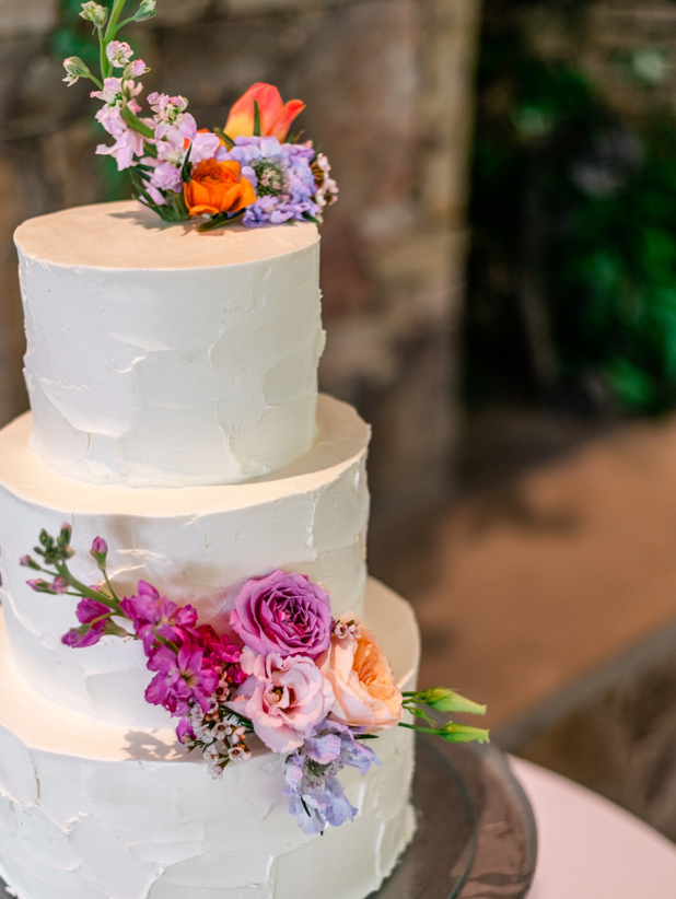 Three-tiered white wedding cake decorated with clusters of colorful fresh flowers, including pink, orange, and purple blooms, displayed on a round table with a blurred stone background.