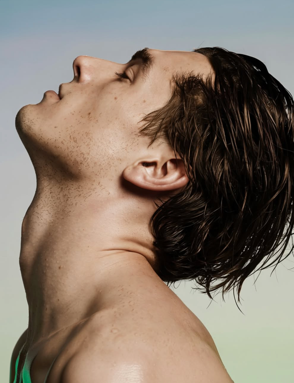 A young man with wet, brown hair tilts his head back and closes his eyes, exposing his neck. The background is softly blurred with gradient colors.