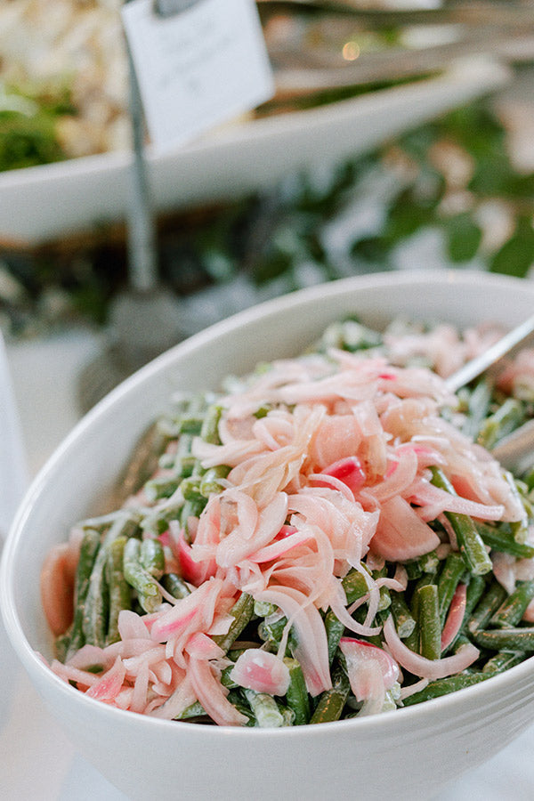 A white bowl filled with green beans topped with thinly sliced pickled red onions, with a serving spoon inside. The background shows another out-of-focus dish and greenery.