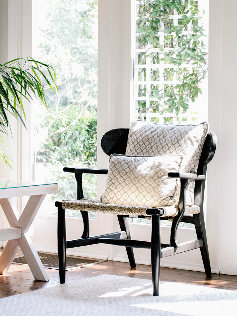 A black wooden chair with two patterned cushions sits by large sunlit windows, surrounded by greenery. A white table and leafy plant are partially visible in the bright, airy room.