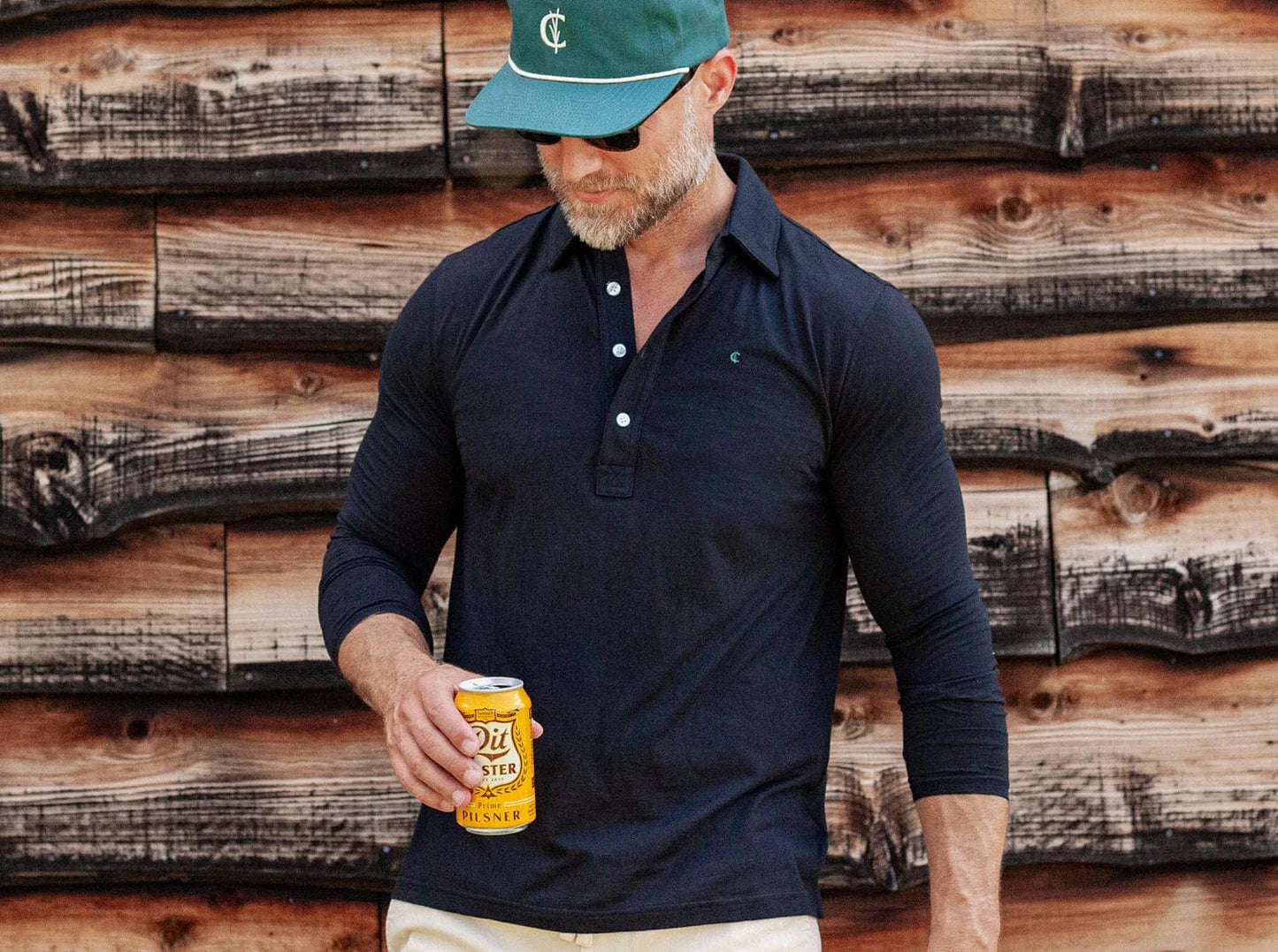 A man wearing a dark long-sleeve polo shirt, beige pants, and a green cap stands in front of a rustic wooden wall, holding a yellow can of beer in his right hand and looking down.
