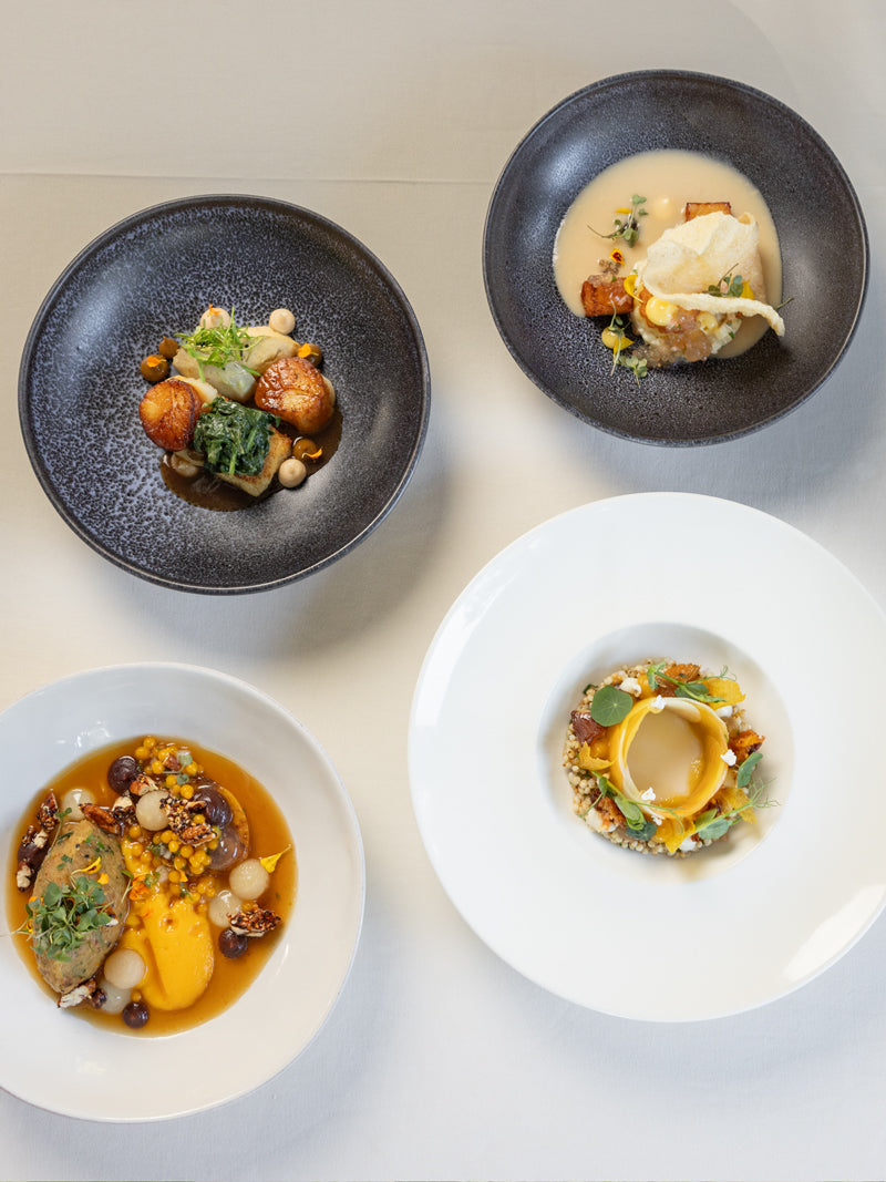 Four elegant plates of gourmet food are artfully arranged on a white tablecloth, each featuring colorful vegetables, sauces, and garnishes in unique presentations on round black and white dishes.