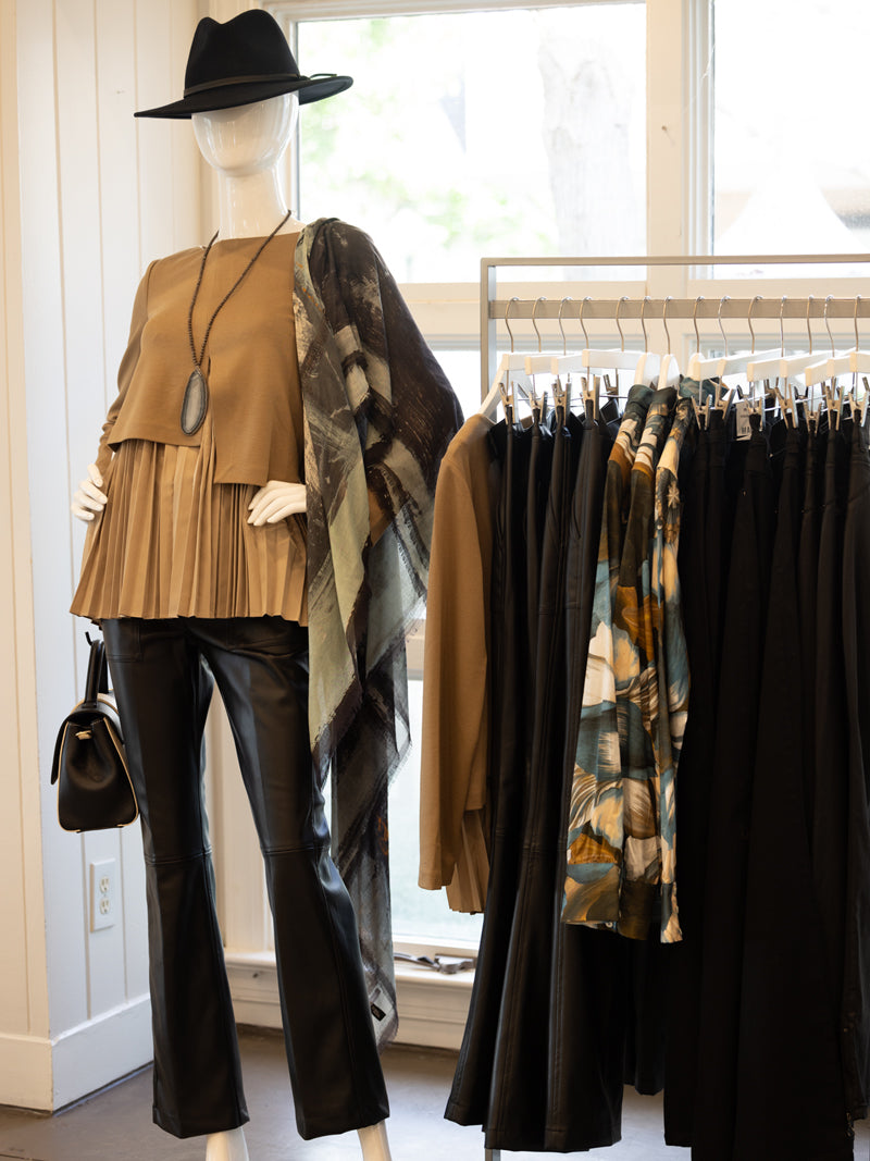 A mannequin dressed in a tan pleated top, black leather pants, a pendant necklace, a black hat, and a shawl stands beside a rack displaying tan tops, black pants, and patterned scarves in a boutique.