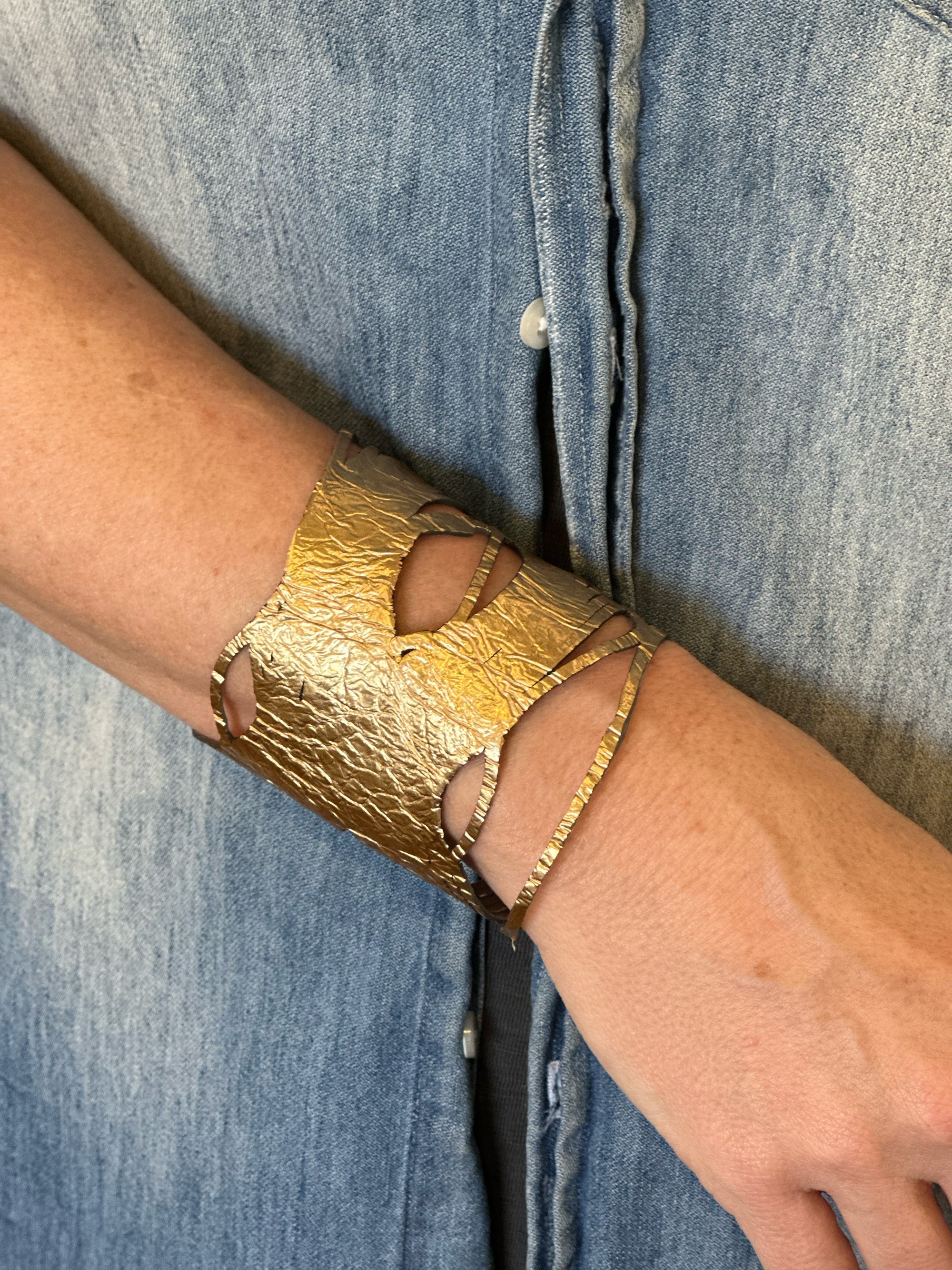 A person wearing a textured, gold-colored cuff bracelet with cutout designs on their wrist, against a background of light blue denim fabric.