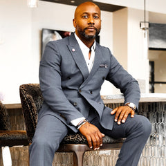 A man in a grey suit sits confidently on a chair with one hand resting on his knee and the other on the chair. He has a trimmed beard and is indoors, with modern decor visible in the background.