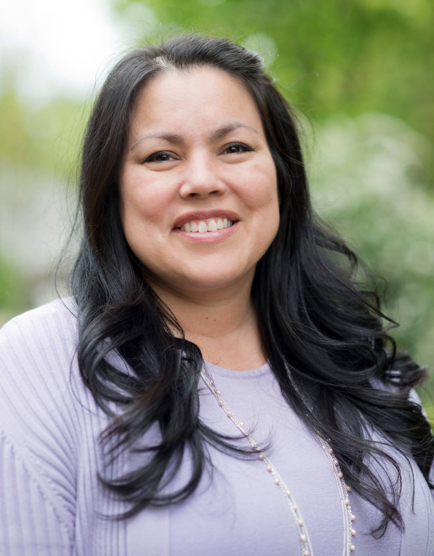 A woman with long dark hair smiles at the camera while standing outdoors, wearing a light purple top and a beaded necklace. The background is blurred with greenery.