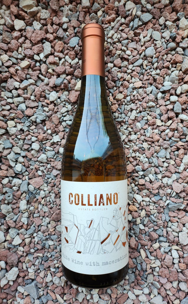 A bottle of Colliano estate bottled wine with a copper-colored cap is placed upright on a surface of small, mixed-color rocks. The label reads "Safe wine with maceration" and features abstract line art.