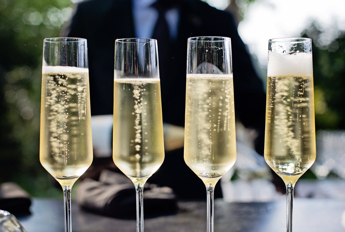 Four tall champagne flutes filled with bubbling champagne sit on a table, with a blurred figure in a dark suit in the background and greenery visible behind them.