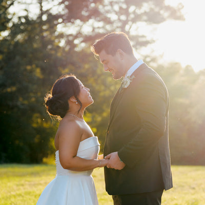 A bride and groom stand outdoors holding hands, gazing at each other and smiling, with sunlight streaming through trees in the background.
