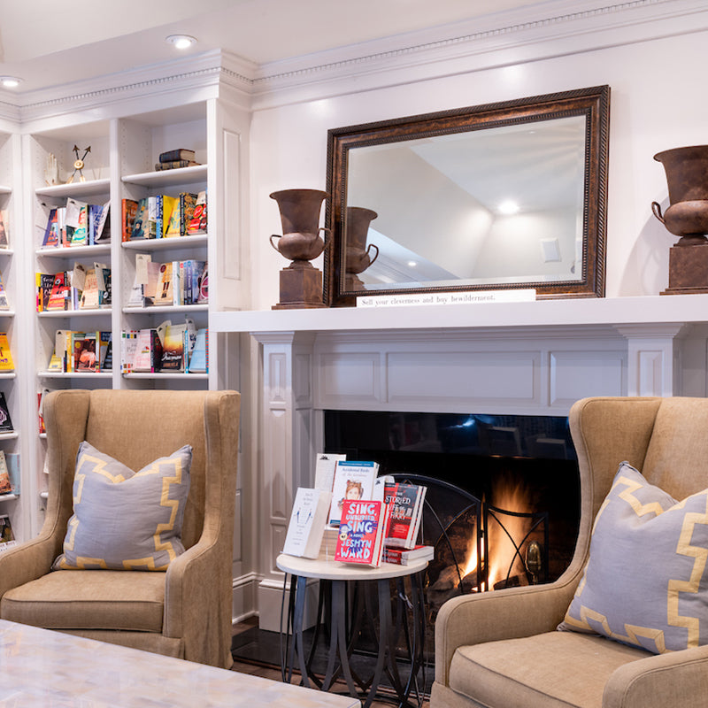 A cozy reading nook features two beige armchairs with gray and yellow pillows, a fireplace with a large mirror above it, bookshelves filled with colorful books, and a stack of books displayed on a small table.