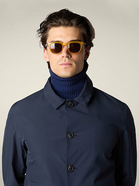 A man wearing brown sunglasses, a navy blue turtleneck, and a dark blue buttoned coat stands against a plain light background, looking straight at the camera with a neutral expression.