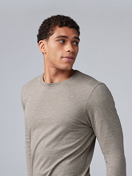 A young man with short curly hair wearing a light gray long-sleeve shirt looks to his left against a plain light gray background.