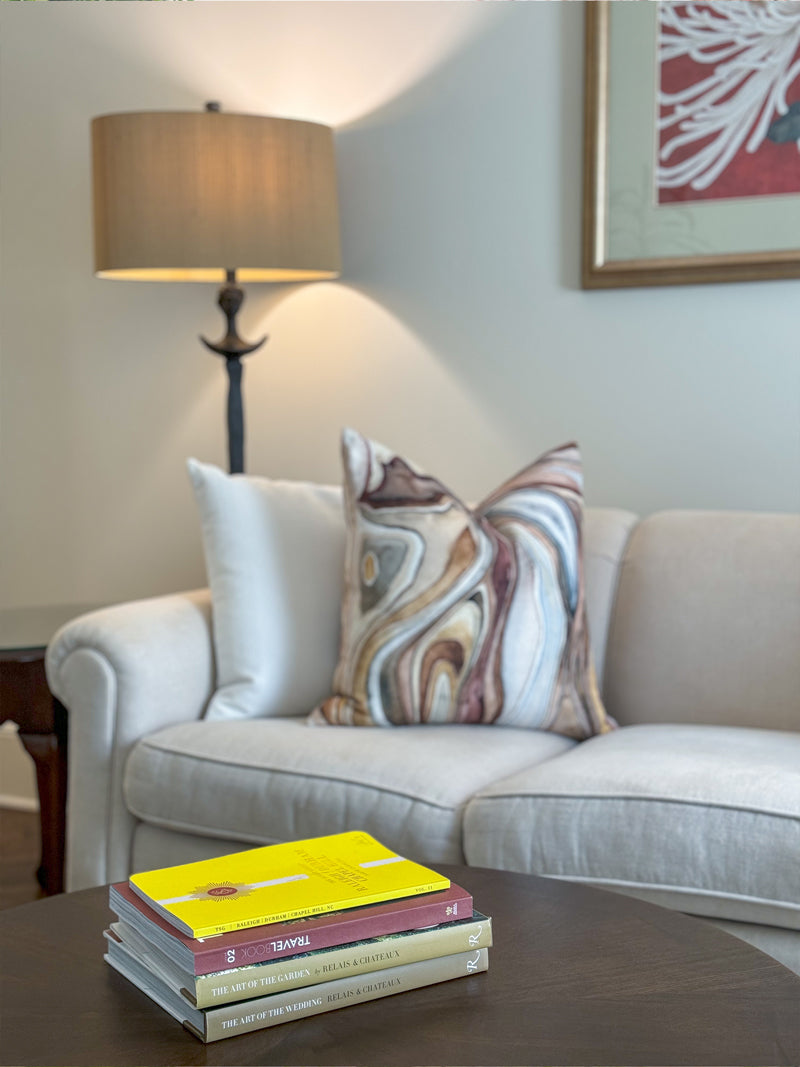 A beige sofa with a patterned pillow sits next to a side table and lamp. In the foreground, a stack of books rests on a dark wooden coffee table. A framed artwork hangs on the wall above the sofa.