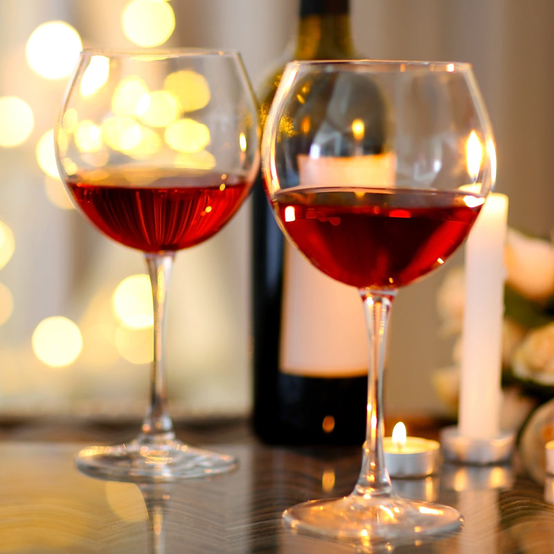 Two glasses of red wine sit on a table with a lit candle, a wine bottle, and soft yellow bokeh lights in the background, creating a warm and romantic atmosphere.