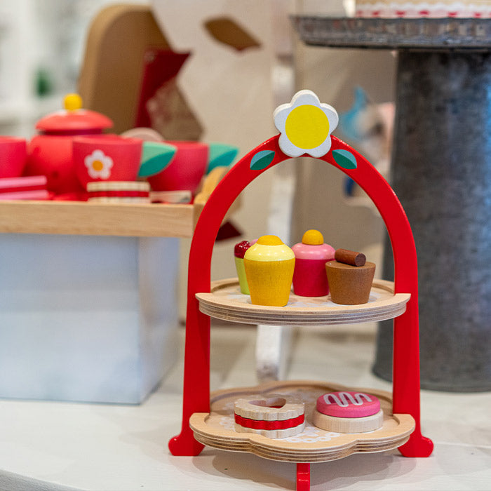 A wooden toy dessert stand with two tiers holds colorful toy pastries and cupcakes. The stand is red with a white and yellow flower detail, and more toy treats are visible in the background.