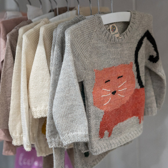 Several children’s sweaters hang on white hangers. The front sweater is gray with a large orange cat design. Other sweaters in pastel shades, such as cream and pink, are visible behind it.
