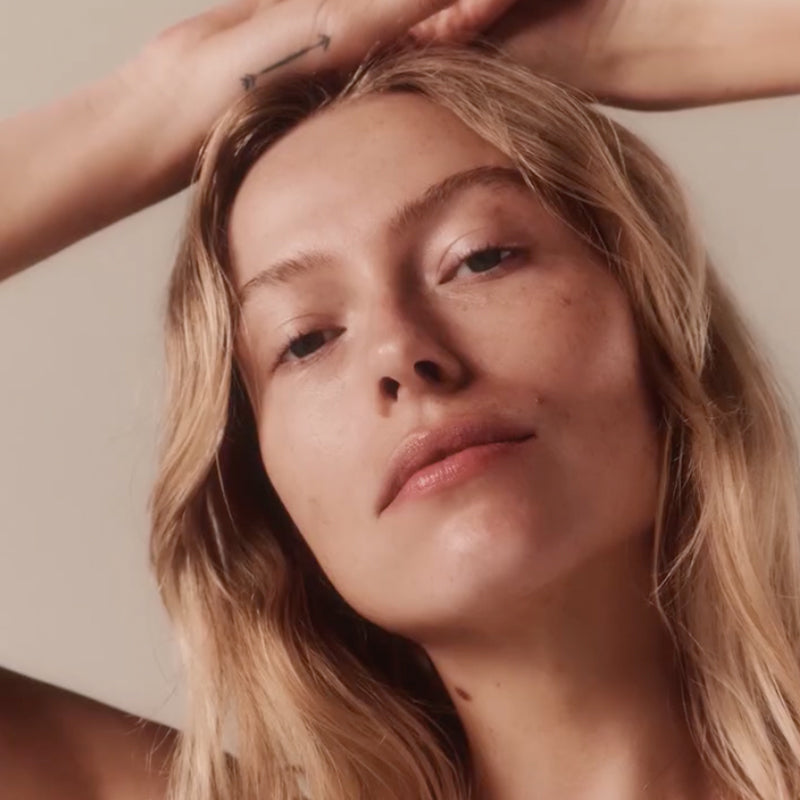 A woman with long blonde hair poses with one arm raised above her head, gazing softly at the camera. Her skin is clear and she has a natural expression on her face. The background is neutral and minimal.