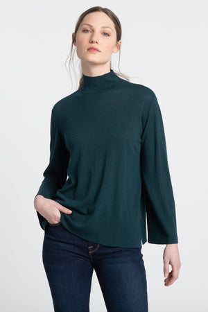 A person with long hair is wearing a KINROSS CASHMERE - EASY MOCK NECK SWEATER in dark green and dark jeans. They have one hand in their pocket and are standing against a plain white background. Their expression is neutral, and they are looking slightly upward.