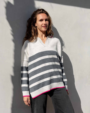 A person with shoulder-length wavy hair is standing against a light-colored wall, wearing the MERSEA - MARINA POLO SWEATER that exudes maritime spirit and dark pants. They are facing the camera with a relaxed expression as shadow patterns fall across the wall behind them.