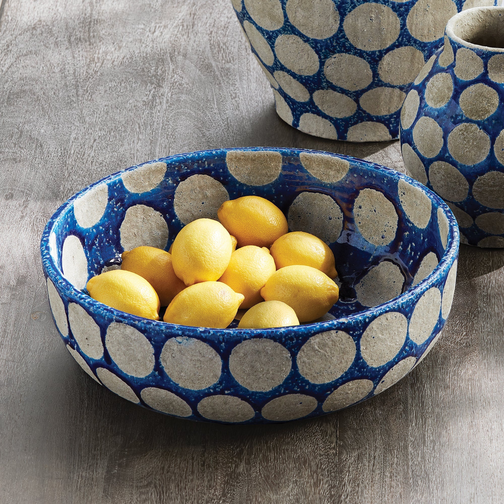 The NAPA HOME AND GARDEN - KILEY BOWL IN BLUE is a cobalt blue ceramic piece with large, textured white polka dots evenly spaced, both inside and out, evoking mid-century pottery. Its wide, rounded design makes it a striking centerpiece for any table.