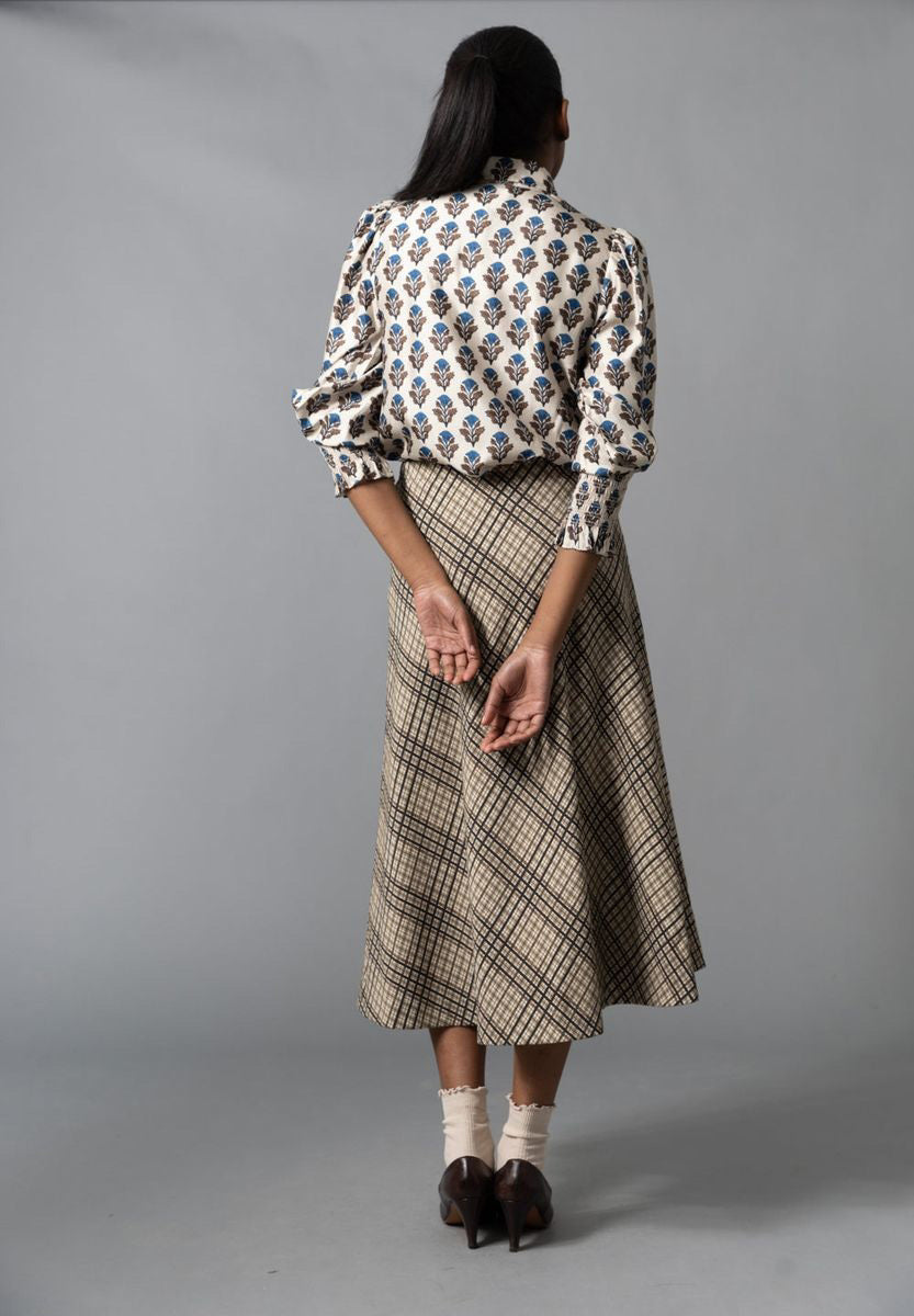 A woman stands in a dimly lit, ornately decorated room, wearing the HOPE FOR FLOWERS - CIRCLE SKIRT IN NATURAL CHECK by HOPE FOR FLOWERS. Warm light from a floor lamp casts shadows on the teal wall behind her.