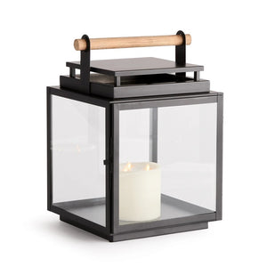 The ADWIN OUTDOOR LANTERN by NAPA HOME AND GARDEN is a black metal and glass outdoor lantern with a square design, featuring an exclusive teak handle on top. Inside the lantern, a lit white candle casts a soft, warm glow.