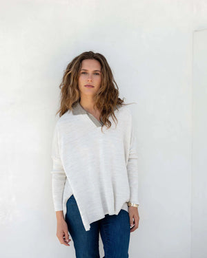 A person with wavy brown hair stands sideways against a plain white background, wearing a light-colored, loose-fitting women's sweater and blue jeans. They glance towards the camera with a neutral expression, showcasing the sporty and stylish appeal of the MERSEA - CATALINA POLO SWEATER by MER SEA.