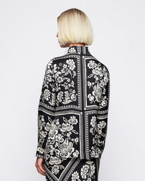 A woman with blonde, chin-length hair is wearing the MIRTO 1956 - BLACK AND WHITE PRINT FLUID BLOUSE. Embracing the intricate flower squares patchwork print of MIRTO fashion, she stands neutrally against a plain white background.