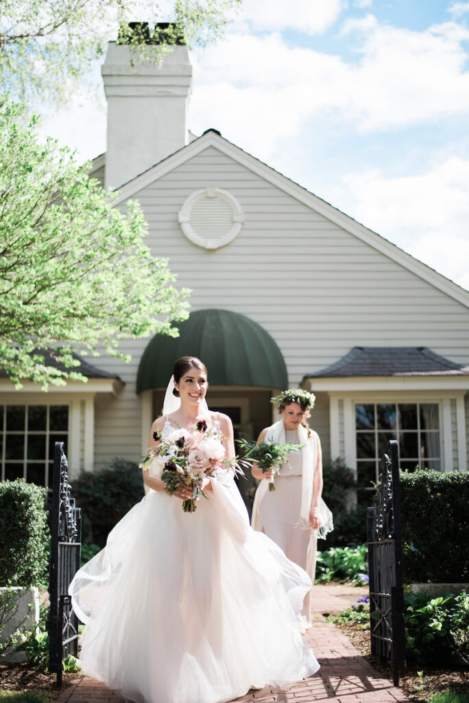 A bride in a flowing white dress holding a bouquet smiles while walking outside a house, followed by a bridesmaid in a light dress and a floral crown. Lush greenery and sunlight frame the scene.