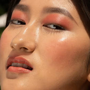 A close-up of a person with curly dark hair, a serious expression, and warm makeup featuring the universally flattering, weightless JASON WU - HOT FLUFF LIP/EYE/CHEEK by JASON WU BEAUTY on their face. They are looking directly at the camera with a neutral background.