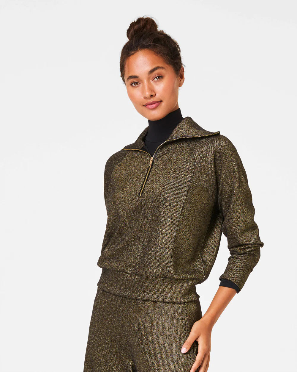 Dressed in the SPANX - AIRESSENTIALS HALF ZIP SHIMMER PULLOVER, a shimmering, gold-toned tracksuit crafted from ultra-soft fabric featuring a quarter-zip top and coordinating pants, a person stands against a simple white background with their hair styled neatly in a bun, radiating a gentle smile.