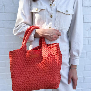 A person wearing a white shirt and dark pants holds the PARKER AND HYDE - WOVEN TOTE BAG with two handles. Their arm, adorned with a gold bracelet, stands out against a plain, light-colored background.