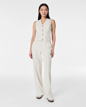 A person with long, straight hair is wearing the SPANX CAREFREE CREPE VEST, a light beige, sleeveless, tailored vest made of easy-care crepe fabric with three buttons and matching trousers from SPANX. Their hands are in their pockets as they pose in front of a plain white background.