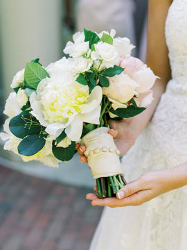 A bride in a white lace dress holds a bouquet of white and pale pink flowers with green leaves, wrapped in a cream-colored ribbon with decorative pearls.