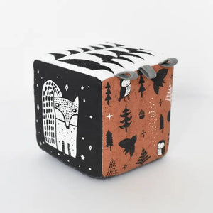 Introducing the WEE GALLERY - WOODLAND SOFT BLOCK by WEE GALLERY, a soft fabric block adorned with a whimsical design perfect for stimulating a baby's developing senses. One side features a black and white squirrel holding a nut, while other sides showcase trees, stars, and owl illustrations in black, white, and brown. Small grey fabric tags protrude from the top for added sensory exploration.