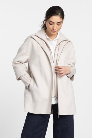 A person stands against a plain background, wearing the KINROSS CASHMERE - KNIT INSERT ZIP MOCK COAT in light beige. The loose-fitting coat features a high-neck collar and is worn over a white shirt. They have dark hair pulled back and are looking down while holding the coat open with both hands. They are also wearing dark pants.