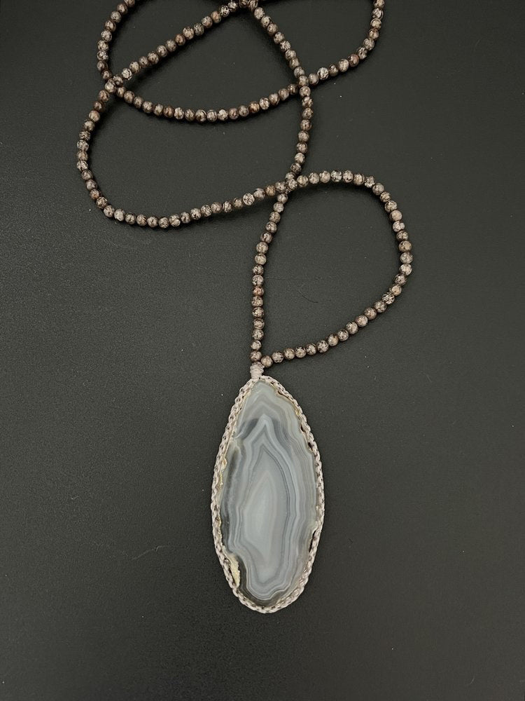 A unique jewelry piece, the ANN LIGHTFOOT Medium Agate Geode Pendant with Snowflake Jasper Strands features a large, polished oval agate geode slice pendant. The stone showcases a distinctive pattern with layered shades of gray, white, and beige that exudes an earthy and natural appeal. The necklace is beautifully displayed against a black background.