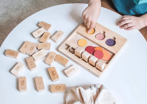 The "LITTLE NUMBERS GAME SET" by LILY & RIVER is a wooden educational toy influenced by Montessori principles. It features number blocks and colored disks on a white surface. The blocks display a simple math equation (2 + 2 = 4), functioning like a children's calculator. In the background, there is a light-colored fabric bag with the text "Lily & River" partially visible.
