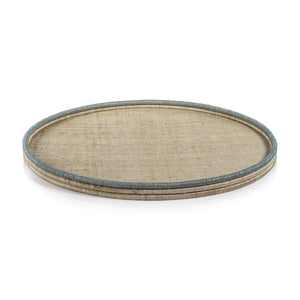 A round, flat **ZODAX** tray with a woven texture sits against a plain white background. The **COILED RATTAN AND ABACA ROUND TRAY**, measuring 24.25 in x 1.5 in, features a beige surface with slightly raised edges encased by a thin, blue border. The design is simple and rustic.