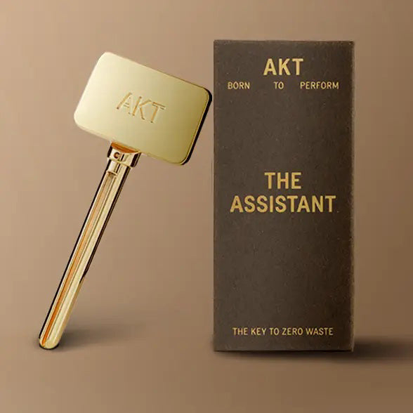 A gold-toned AKT LONDON &quot;THE ASSISTANT KEY TOOL&quot; is shown beside a brown box with gold text, &quot;AKT Born to Perform THE ASSISTANT The Key to Zero Waste,&quot; emphasizing eco-friendly, zero-waste living.