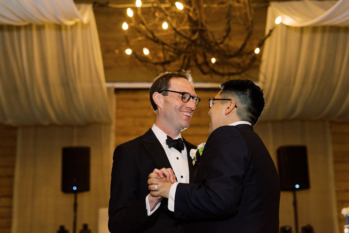 Two men in tuxedos smile and hold hands while dancing together under a chandelier, in a warmly lit venue with draped fabric on the ceiling.